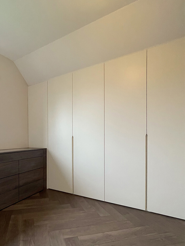 Dressing sur mesure à Luxembourg avec des armoires blanches élégantes, conçues pour s'adapter parfaitement aux pentes du plafond et occupant deux murs de la pièce pour une organisation optimale des vêtements